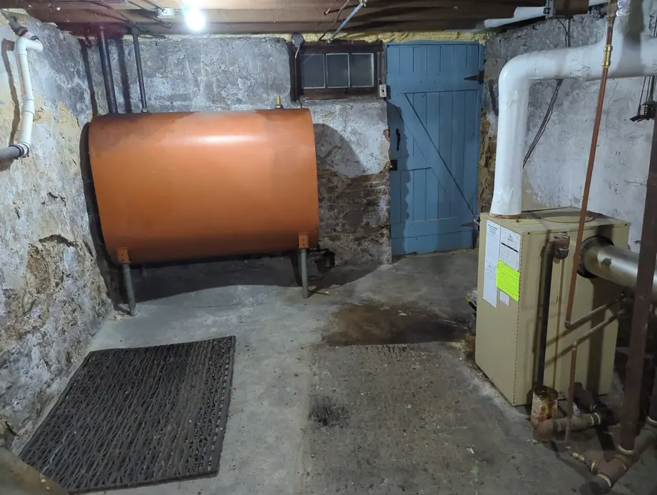 Stone basement before the replacement, with a fuel-oil tank on steel legs and the old boiler beside a blue cellar door